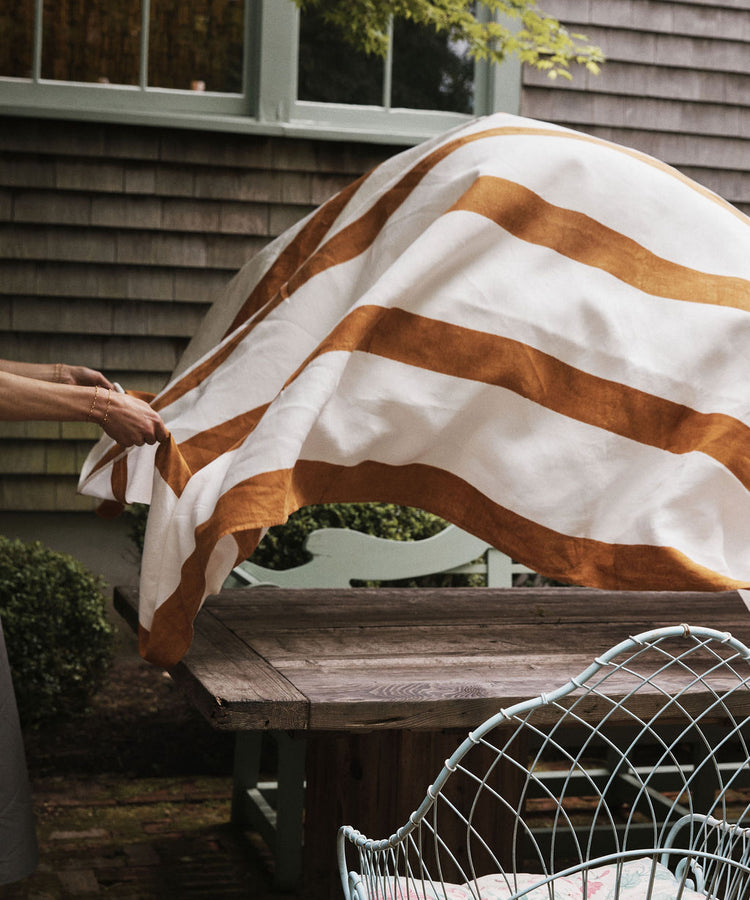 A person spreads the Riga Tablecloth, featuring hand-printed wide brown stripes, over a wooden outdoor table next to a metal chair and house siding.