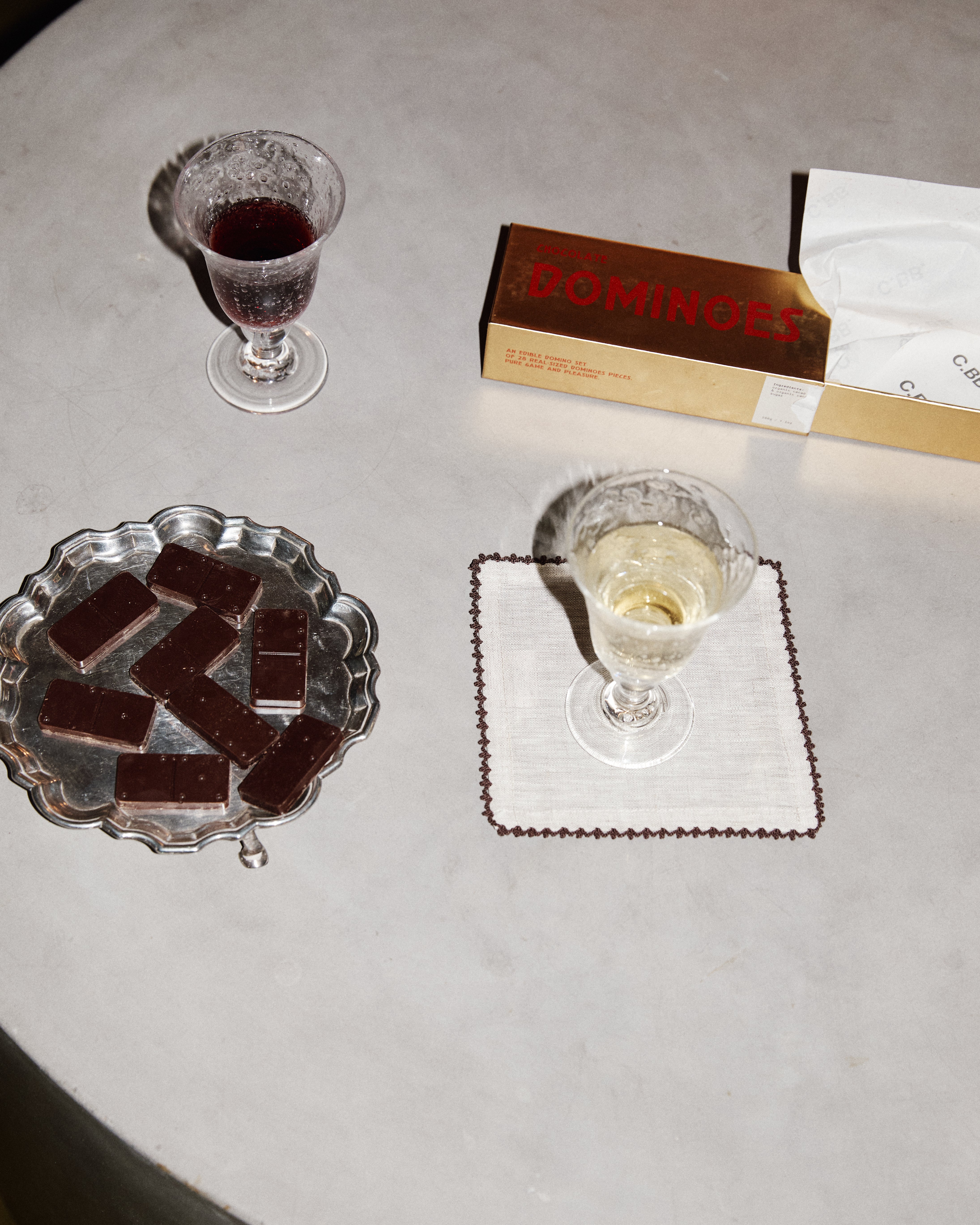 Two glasses of wine, a plate of chocolate bars, and a box of Dominoes on a gray surface.
