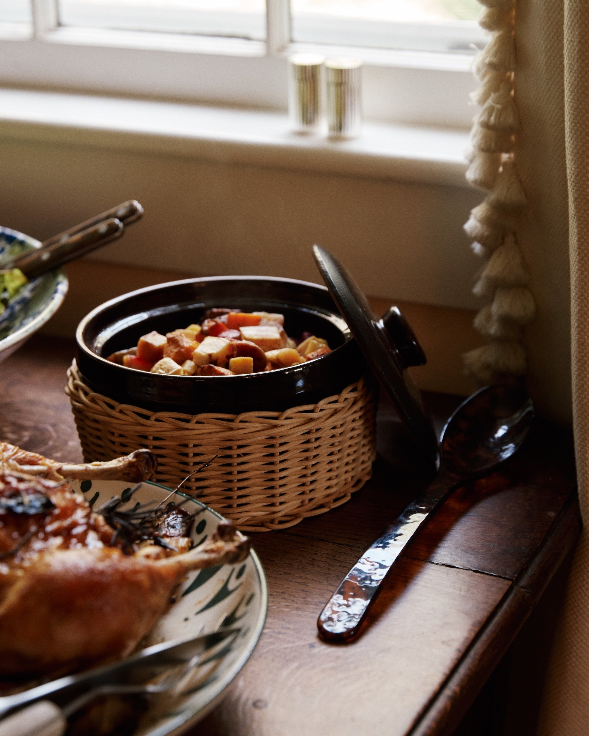 An Amany Casserole filled with diced vegetables in a ceramic dish with wicker sleeve rests on a wooden table next to a serving spoon and roasted meat.