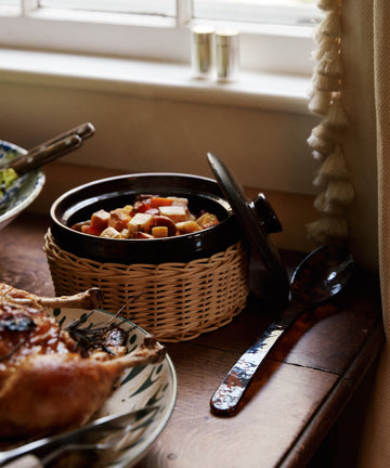 An Amany Casserole filled with diced vegetables in a ceramic dish with wicker sleeve rests on a wooden table next to a serving spoon and roasted meat.