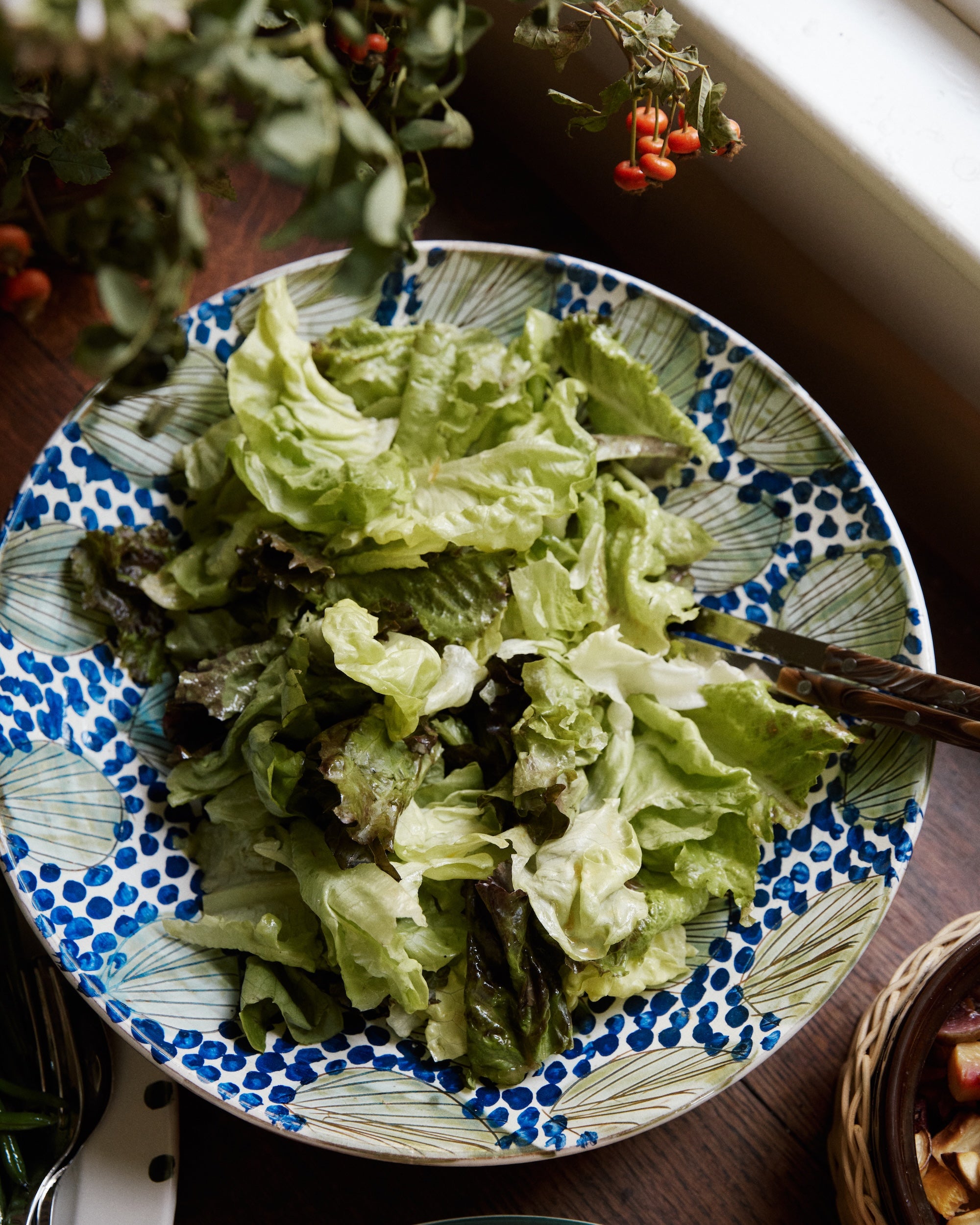 A mix of green and dark leafy lettuce in a Green Leaves Serving Bowl on a wooden table near a window with plants in the background.