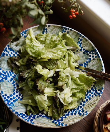 A mix of green and dark leafy lettuce in a Green Leaves Serving Bowl on a wooden table near a window with plants in the background.