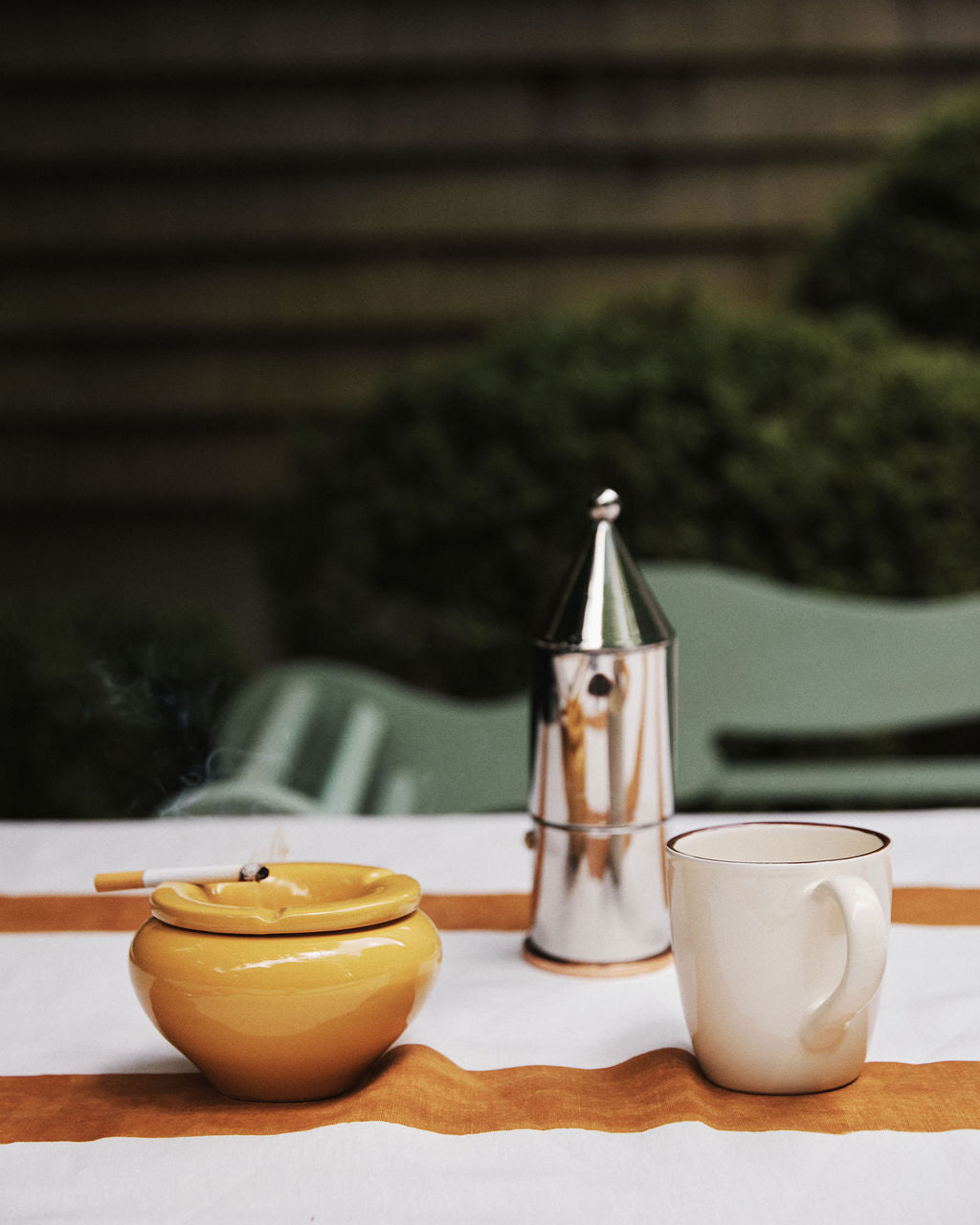 The Mix & Match Ashtray, handmade in Italy with two-tone glazes, sits on an outdoor table with a lit cigarette, a silver moka pot, and a white mug atop an orange-striped cloth.