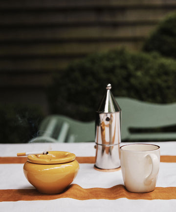 The Mix & Match Ashtray, handmade in Italy with two-tone glazes, sits on an outdoor table with a lit cigarette, a silver moka pot, and a white mug atop an orange-striped cloth.