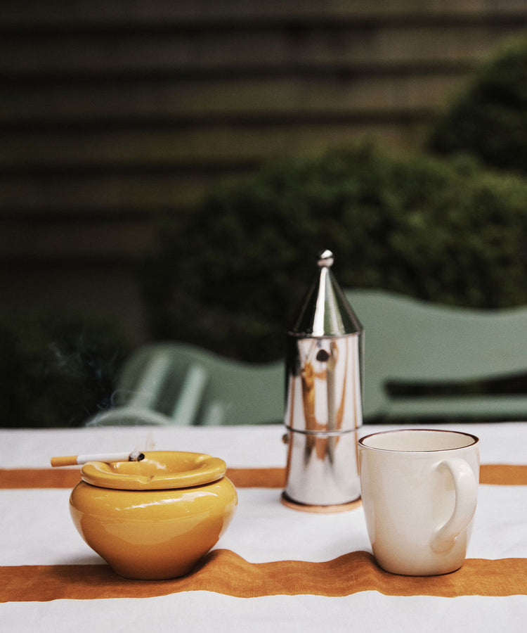 The Mix & Match Ashtray, handmade in Italy with two-tone glazes, sits on an outdoor table with a lit cigarette, a silver moka pot, and a white mug atop an orange-striped cloth.