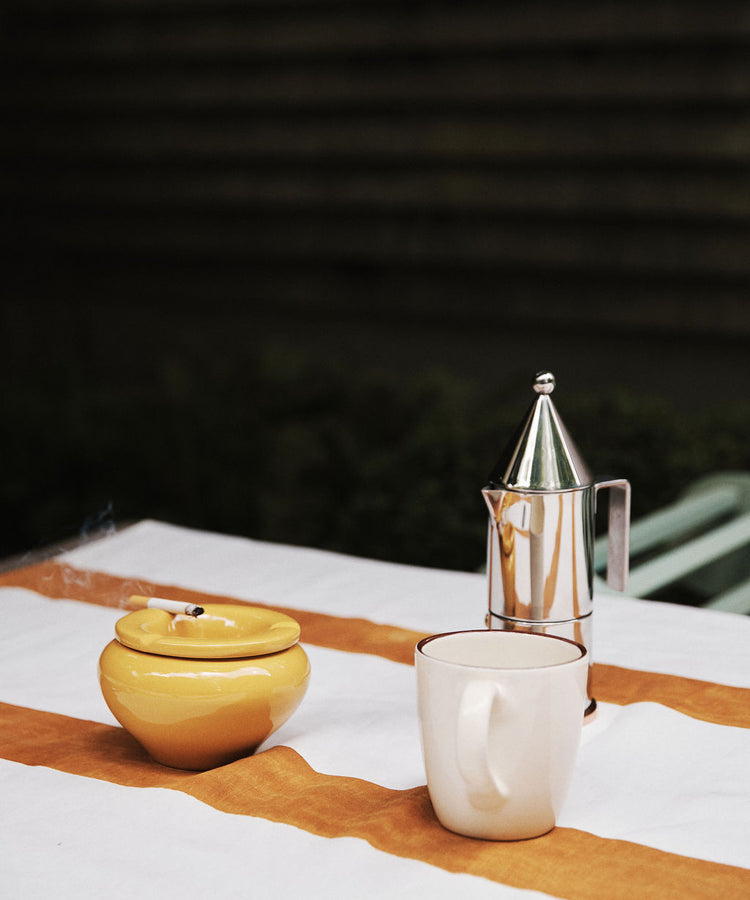 A white mug, a yellow ashtray with a lit cigarette, and the La Conica Espresso Coffee Maker are arranged on a table covered with a white and orange striped cloth.