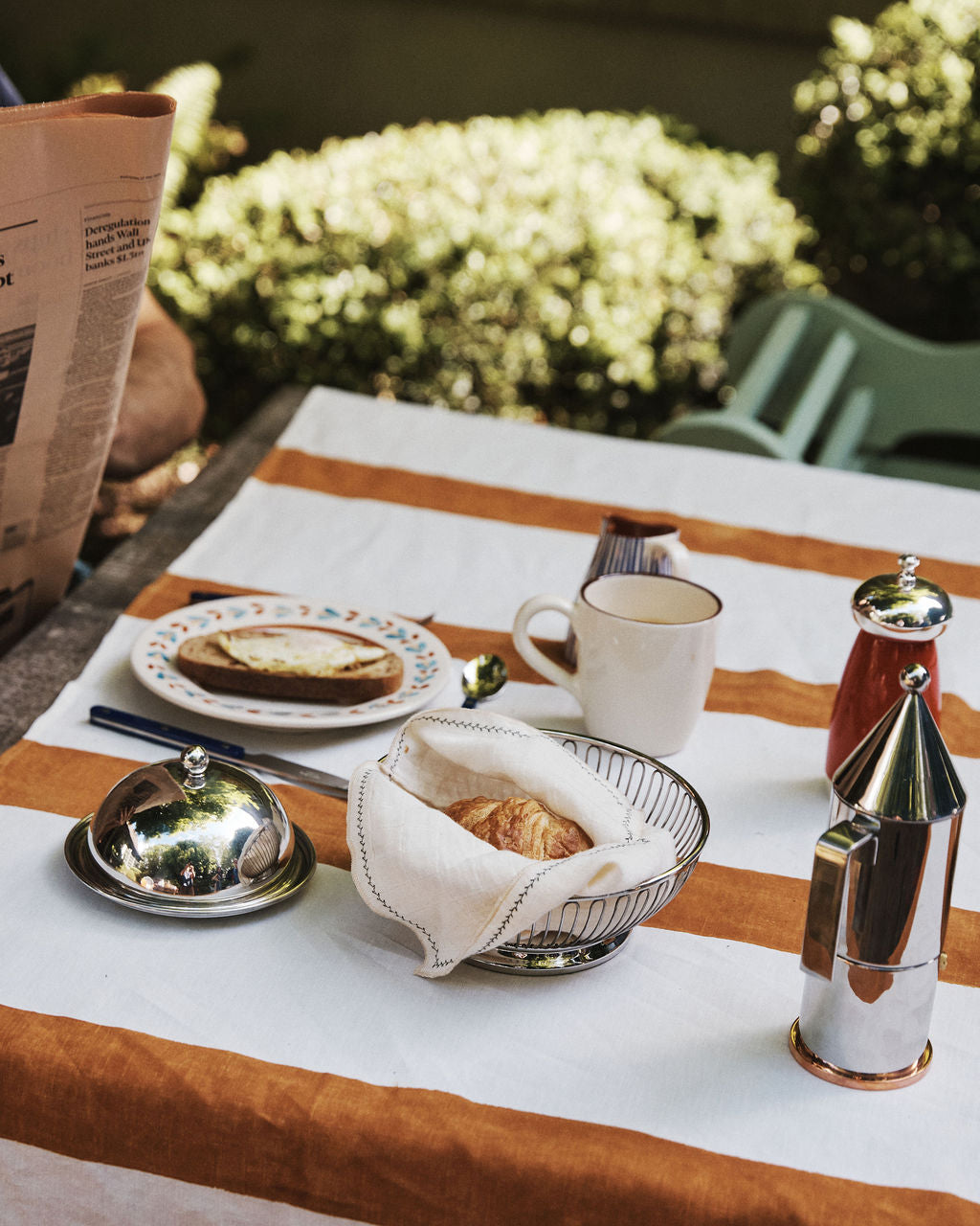 A breakfast table set outdoors on a striped tablecloth features a croissant in the Oval Bread Basket, accompanied by a cup, a plate with bread and cheese, a coffee pot, a creamer, and a pepper grinder.