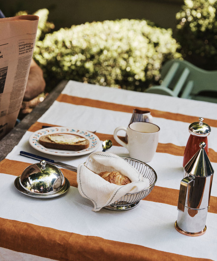 A breakfast table set outdoors on a striped tablecloth features a croissant in the Oval Bread Basket, accompanied by a cup, a plate with bread and cheese, a coffee pot, a creamer, and a pepper grinder.