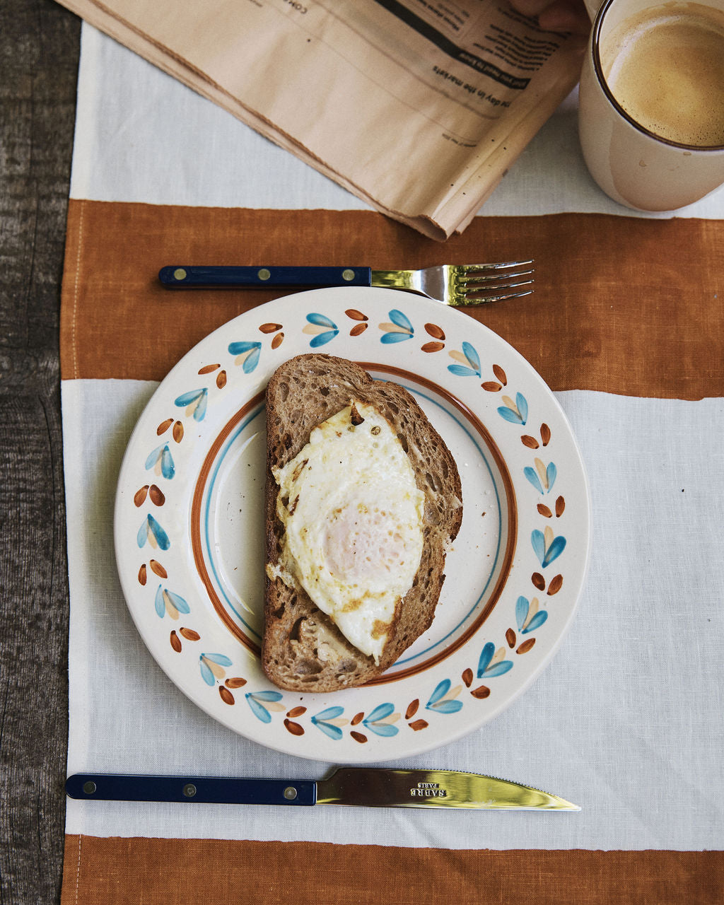 A slice of bread with a fried egg rests on an Ottoline Small Plate, set of 4, accompanied by a fork, knife, cup of coffee, and a folded newspaper atop a striped tablecloth.