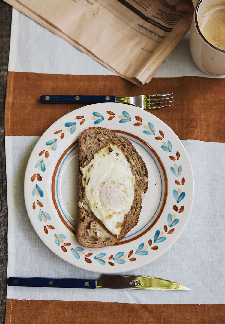 A slice of bread with a fried egg rests on an Ottoline Small Plate, set of 4, accompanied by a fork, knife, cup of coffee, and a folded newspaper atop a striped tablecloth.