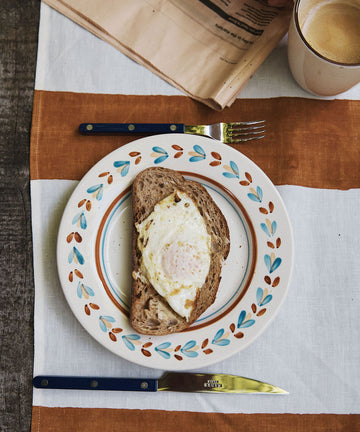 A slice of bread with a fried egg rests on an Ottoline Small Plate, set of 4, accompanied by a fork, knife, cup of coffee, and a folded newspaper atop a striped tablecloth.