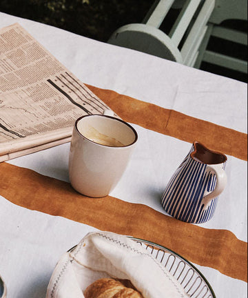 A white mug, the handmade Natalia Milk Jug for the breakfast table, a croissant in a basket, and a folded newspaper rest on a table with a white and orange tablecloth.