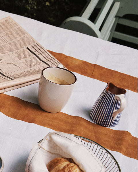 A white mug, the handmade Natalia Milk Jug for the breakfast table, a croissant in a basket, and a folded newspaper rest on a table with a white and orange tablecloth.