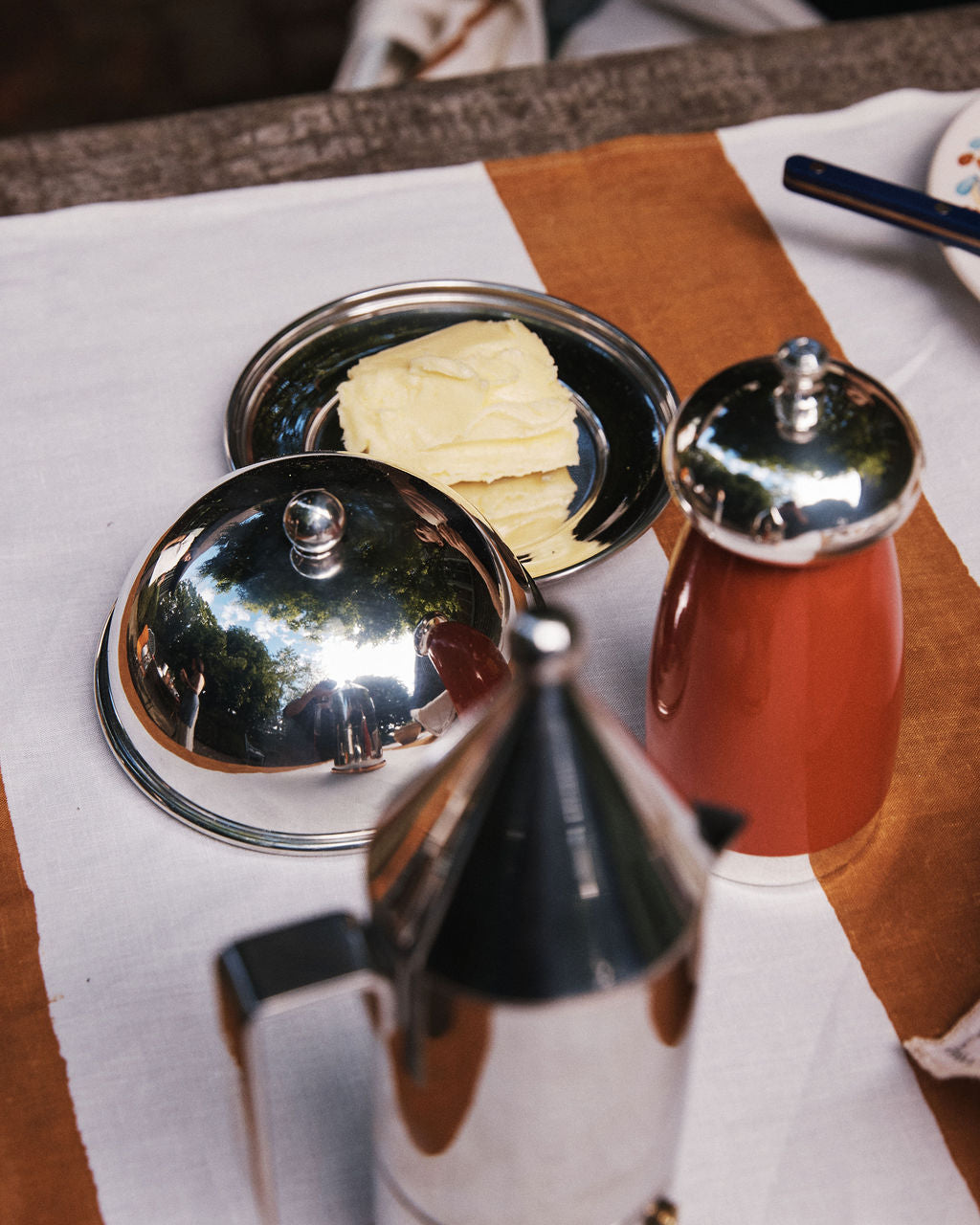 Rectangular slices of butter rest on the elegant Nell Butter Dish with a lid, set beside a red container and a metallic coffee pot, all arranged on a white and brown tablecloth.