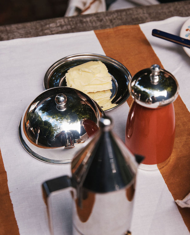 Rectangular slices of butter rest on the elegant Nell Butter Dish with a lid, set beside a red container and a metallic coffee pot, all arranged on a white and brown tablecloth.