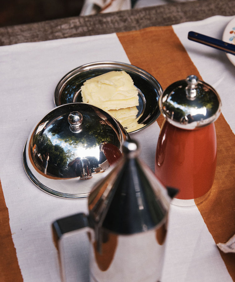 Rectangular slices of butter rest on the elegant Nell Butter Dish with a lid, set beside a red container and a metallic coffee pot, all arranged on a white and brown tablecloth.
