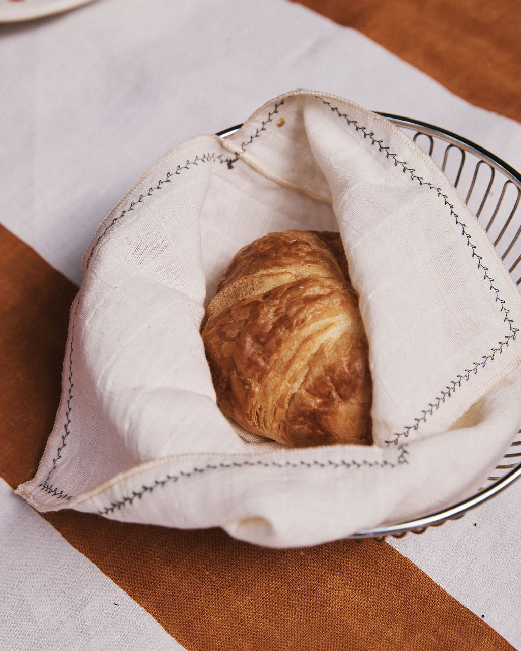 A Filial Bread Bento, Set of 2, wrapped in a white cloth napkin rests in a wire basket atop a table dressed with a white and brown striped tablecloth, blending picnic casualness with the refined elegance of Japanese linens.