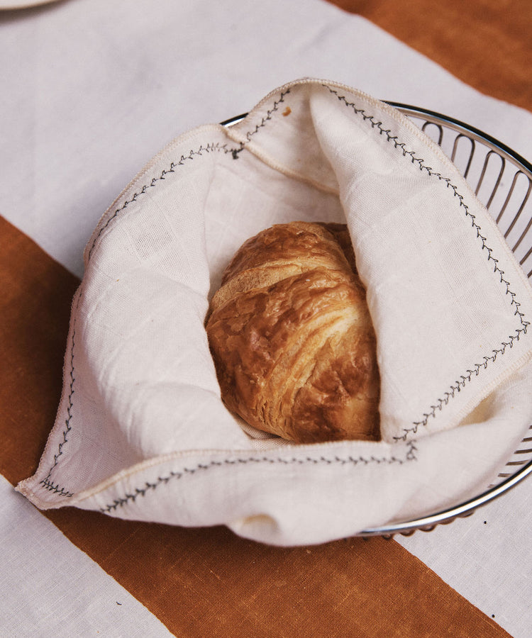 A Filial Bread Bento, Set of 2, wrapped in a white cloth napkin rests in a wire basket atop a table dressed with a white and brown striped tablecloth, blending picnic casualness with the refined elegance of Japanese linens.