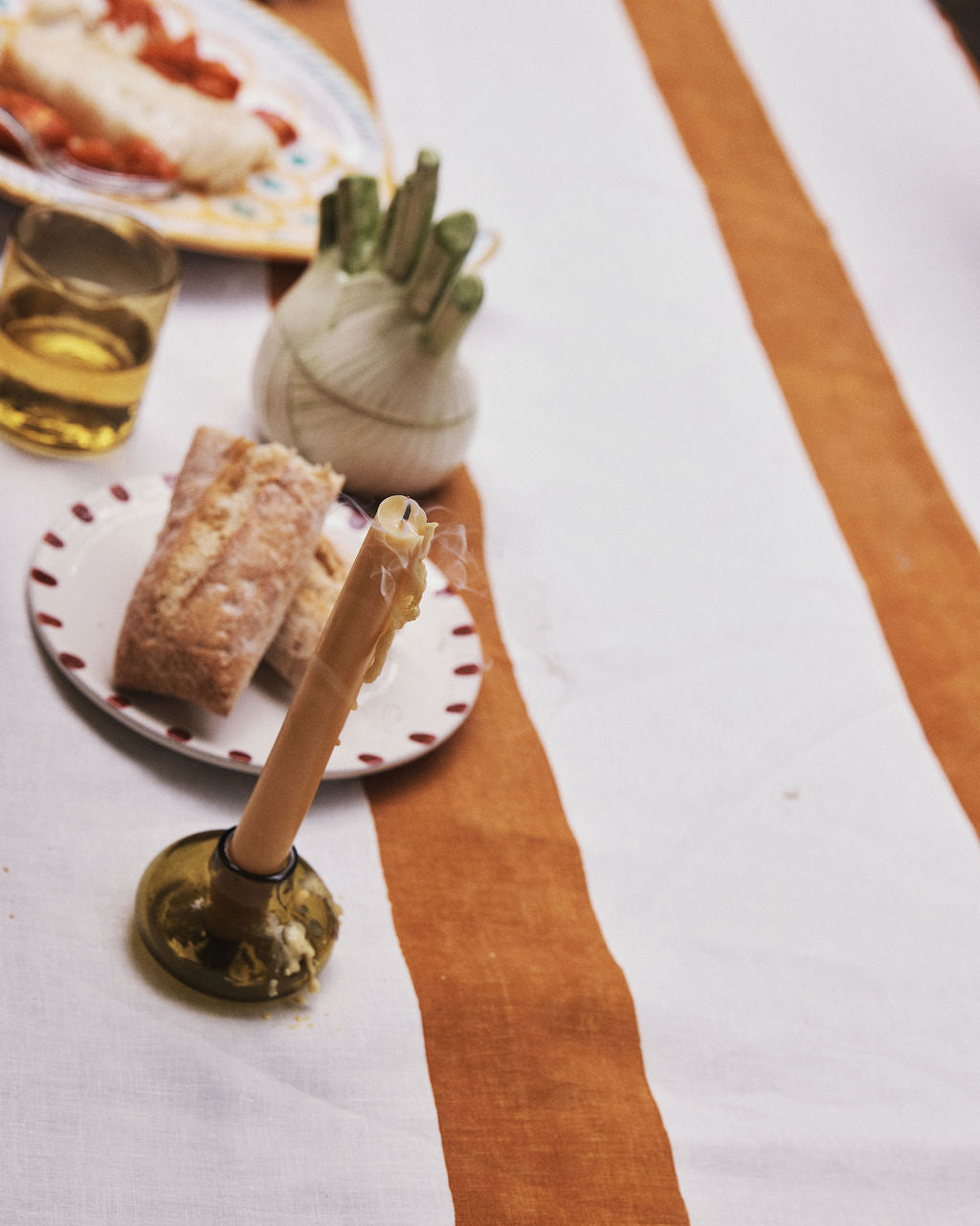 A striped tablecloth with a candle set in our Lucien bubble candleholder, Large—a handblown piece made from recycled glass in Morocco—adds an artisan touch to your dining setting. Nearby is a glass of drink, bread on a plate, and a vegetable holder filled with fresh green vegetables.