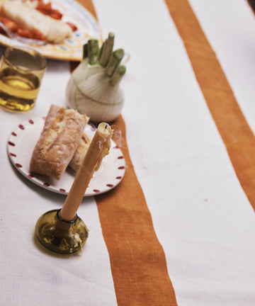 A striped tablecloth with a candle set in our Lucien bubble candleholder, Large—a handblown piece made from recycled glass in Morocco—adds an artisan touch to your dining setting. Nearby is a glass of drink, bread on a plate, and a vegetable holder filled with fresh green vegetables.