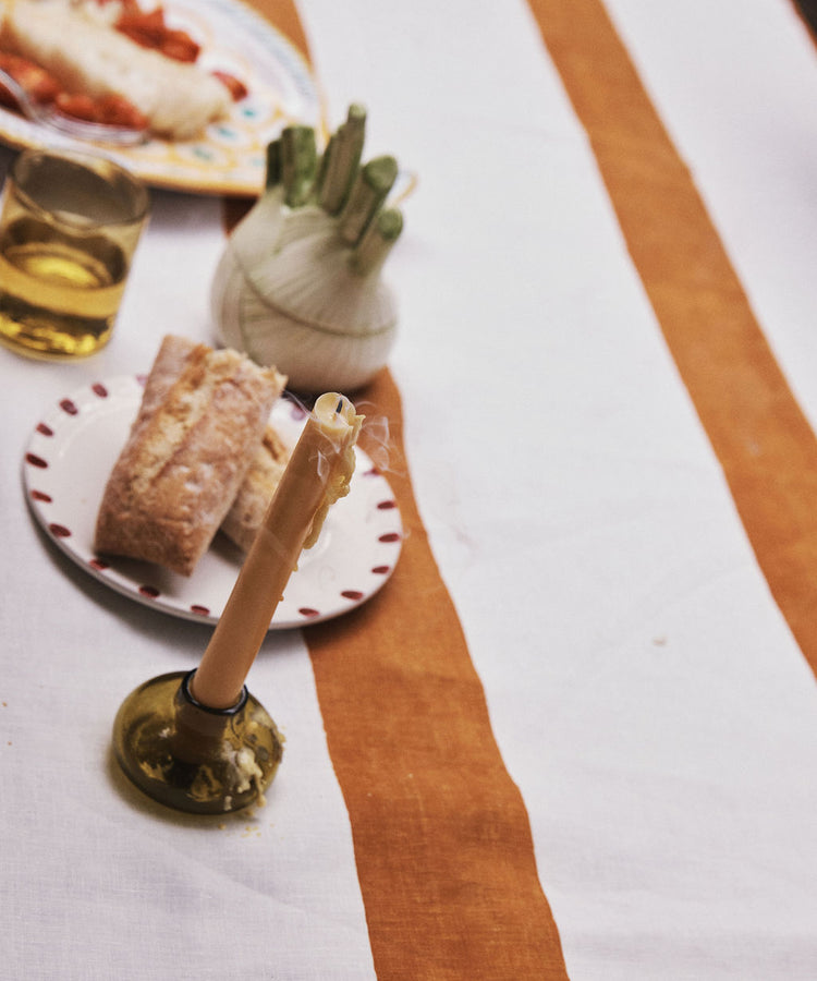 A striped tablecloth with a candle set in our Lucien bubble candleholder, Large—a handblown piece made from recycled glass in Morocco—adds an artisan touch to your dining setting. Nearby is a glass of drink, bread on a plate, and a vegetable holder filled with fresh green vegetables.
