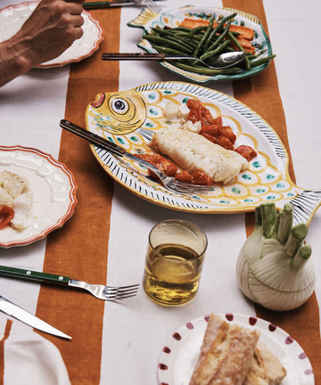 A meal featuring fish fillet and tomatoes is served on a playful Ricardo Serving Platter shaped like a fish, accompanied by green beans, carrots, bread, a drink, and fennel.