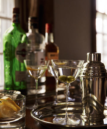 A perfect martini in a Tina Martini Glass, Set of 4 sits with olives, a shaker, and bottles of liquor on a tray by a sunlit window.