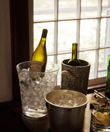 Two wine bottles, one chilled in a Lauren Champagne Bucket of French glassware, sit by a window with ice buckets and a small dish of lemon slices on the table.