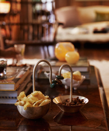 The Astrid Aperitivo Set with chips and nuts sits on a wooden table alongside books, glasses, and candles; a sofa is blurred in the background.
