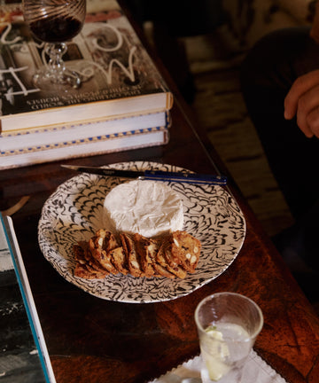 The Celine Serving Platter displays brie and fruit and nut crackers on a wooden table, next to a pen, books, and a glass of wine.