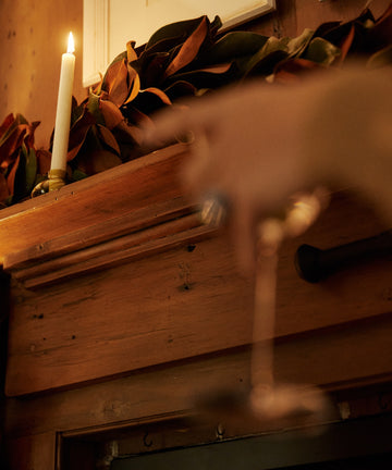A lit candle in the Lucien bubble candleholder, Large, sits on a wooden mantel with autumn leaves; a hand holding a wine glass is blurred in the foreground.