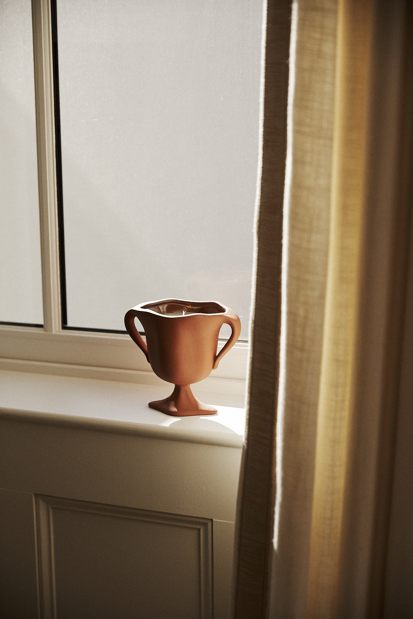 A Herbarium Candle sits on a windowsill, sunlight streaming through a cream-colored curtain and filling the air with gentle botanical scents.