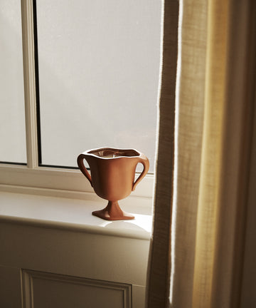 A Herbarium Candle sits on a windowsill, sunlight streaming through a cream-colored curtain and filling the air with gentle botanical scents.