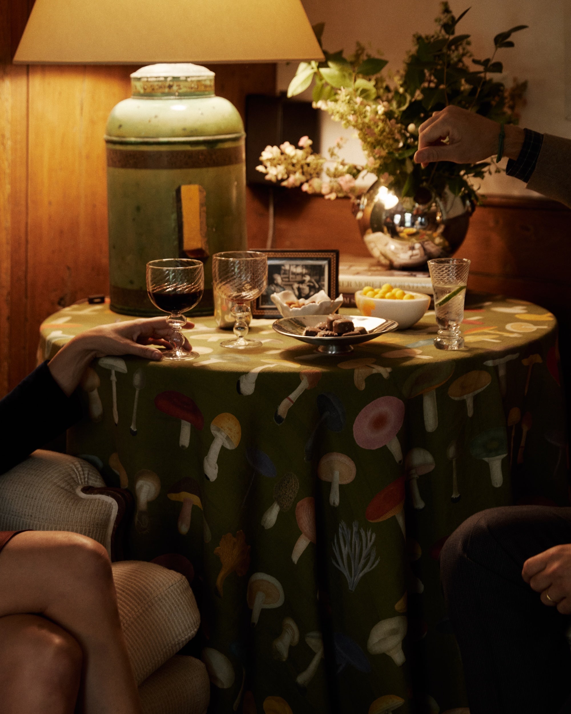 A small table covered with the John Derian Fungi Round Tablecloth holds glasses, snacks, a photo frame, and a lamp. Two people’s arms are visible—one pouring a drink.