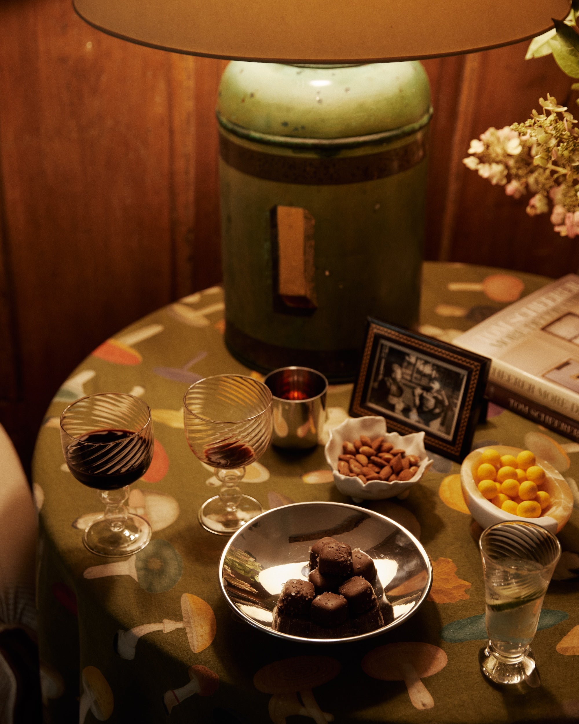 A lamp on a round table with two glasses of red wine, an Elena Bowl of almonds, chocolates, yellow candies, a framed photo, a book, and flowers.