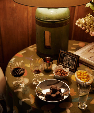 A lamp on a round table with two glasses of red wine, an Elena Bowl of almonds, chocolates, yellow candies, a framed photo, a book, and flowers.