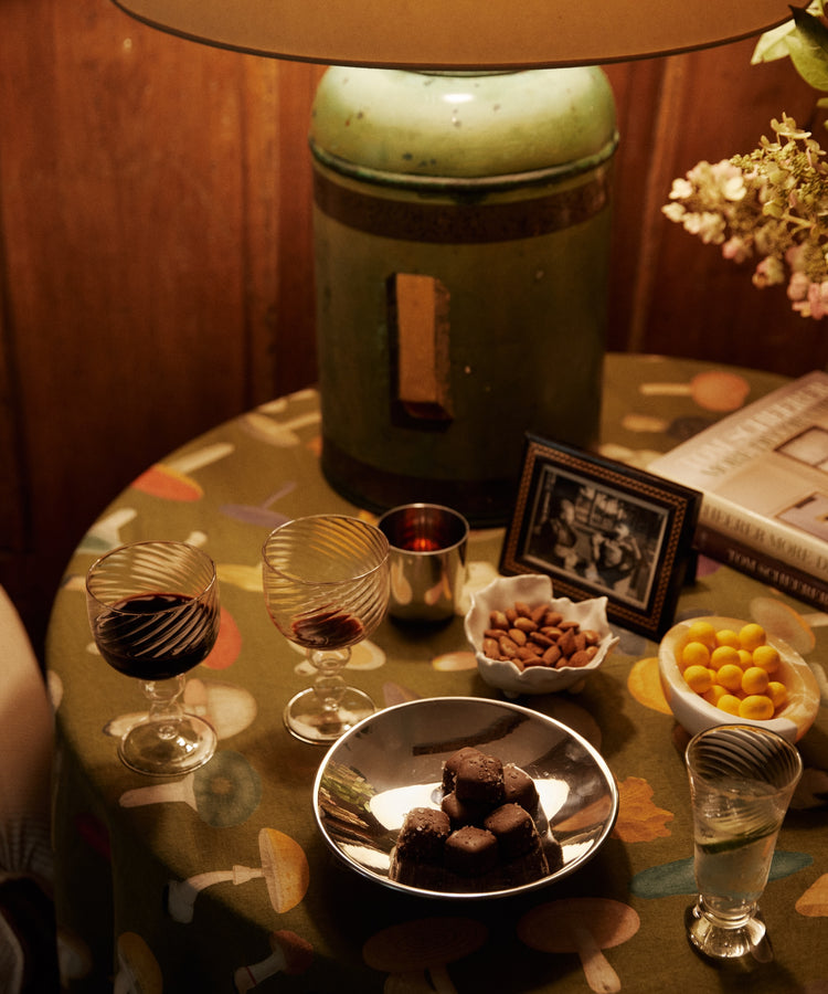A lamp on a round table with two glasses of red wine, an Elena Bowl of almonds, chocolates, yellow candies, a framed photo, a book, and flowers.