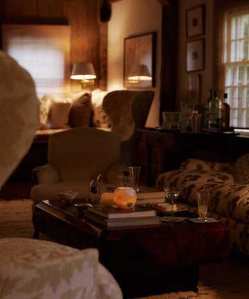 A cozy living room features patterned armchairs, a wooden coffee table with books, fruit, and the Akila Candleholder for unique decor under warm lamps and window light.