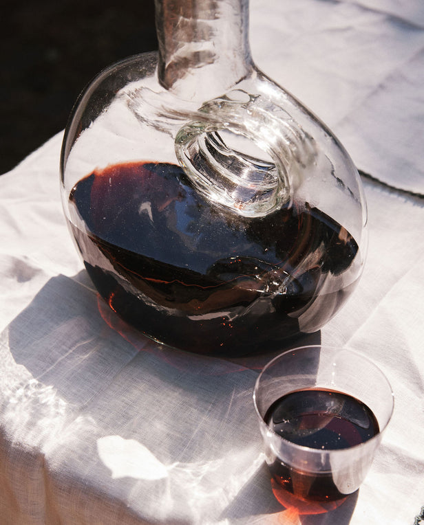 An Alma Decanter, hand-blown in Spain and filled with red wine, sits next to a plastic cup of red wine on a white tablecloth in sunlight.
