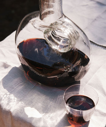 An Alma Decanter, hand-blown in Spain and filled with red wine, sits next to a plastic cup of red wine on a white tablecloth in sunlight.