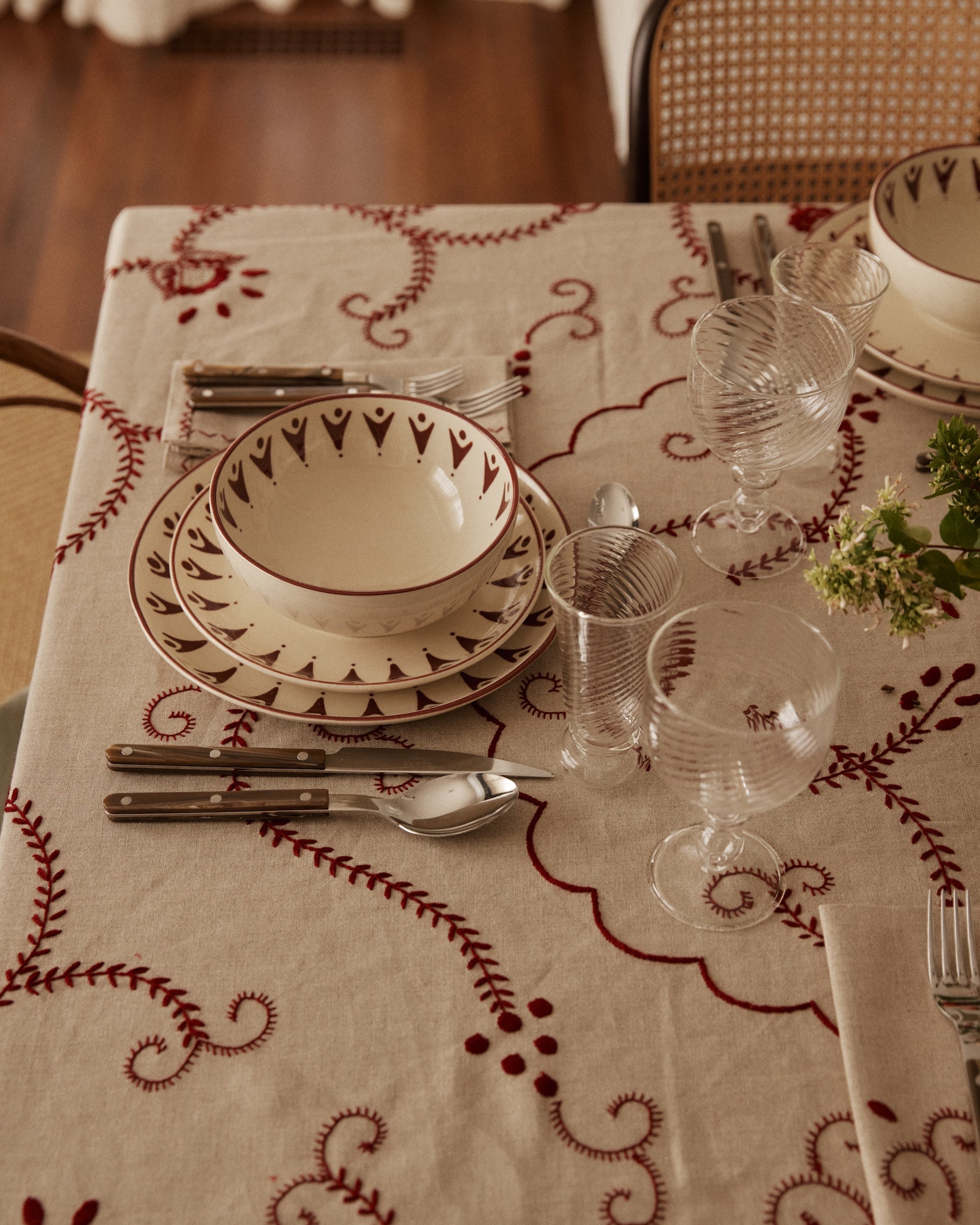 A table set with patterned plates, bowls, cutlery, ribbed glasses, and the Inez Tablecloth highlights traditional designs with a small green flower arrangement.