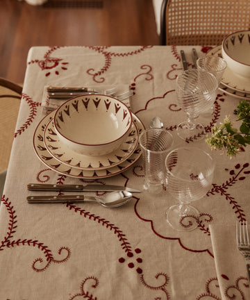 A table set with patterned plates, bowls, cutlery, ribbed glasses, and the Inez Tablecloth highlights traditional designs with a small green flower arrangement.