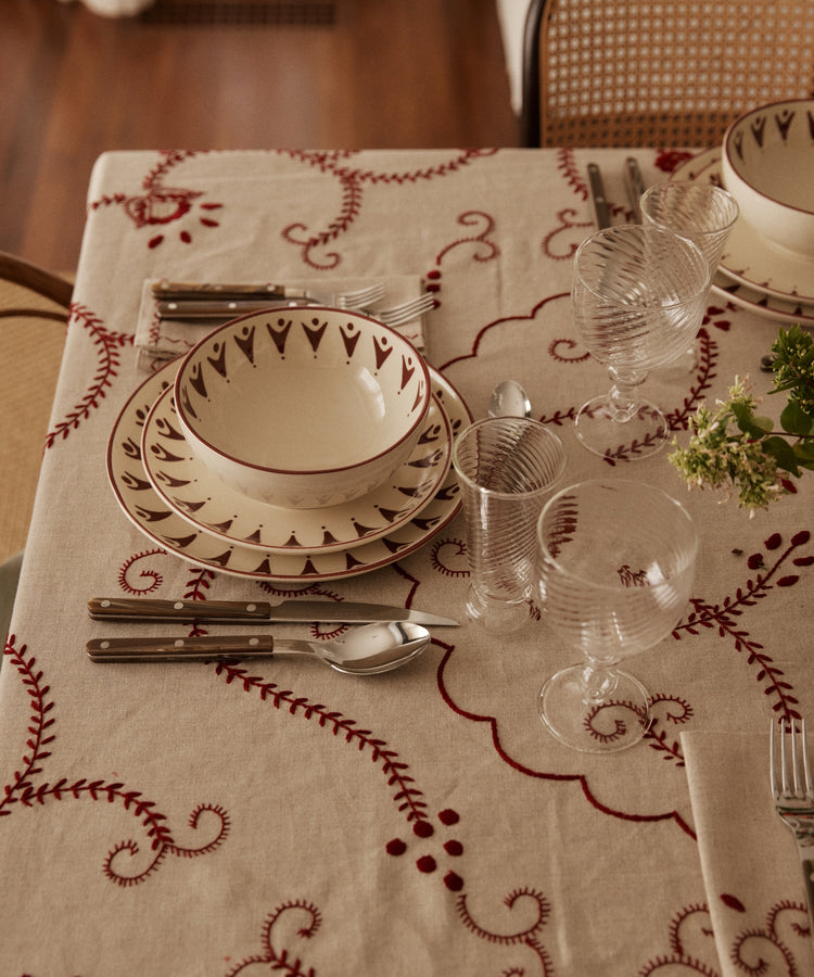 A table set with patterned plates, bowls, cutlery, ribbed glasses, and the Inez Tablecloth highlights traditional designs with a small green flower arrangement.