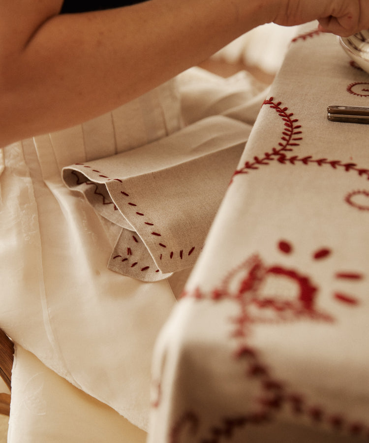 A person rests their arm on a table set with a raw linen tablecloth and the Inez Napkin, Set of 6, holding a plate; only their arm and torso are shown.