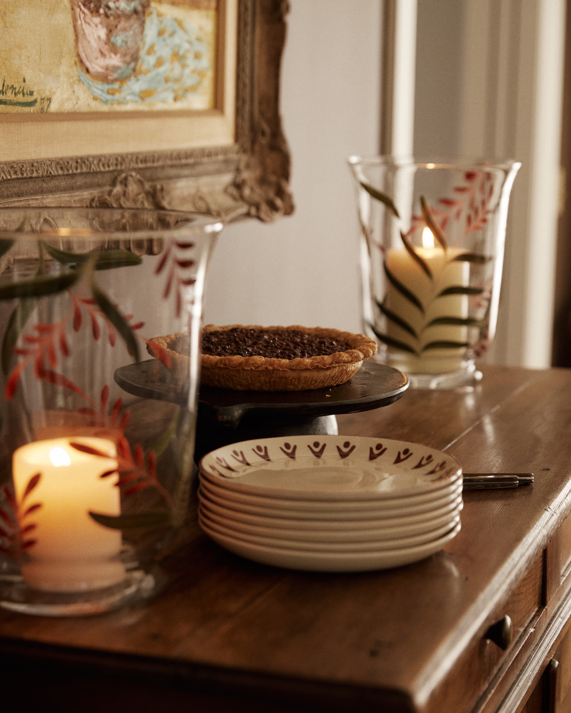 A pecan pie on a cake stand, Vito Small Plate, Set of 4, a fork, and glass candle holders with leaf designs sit on a wooden sideboard.