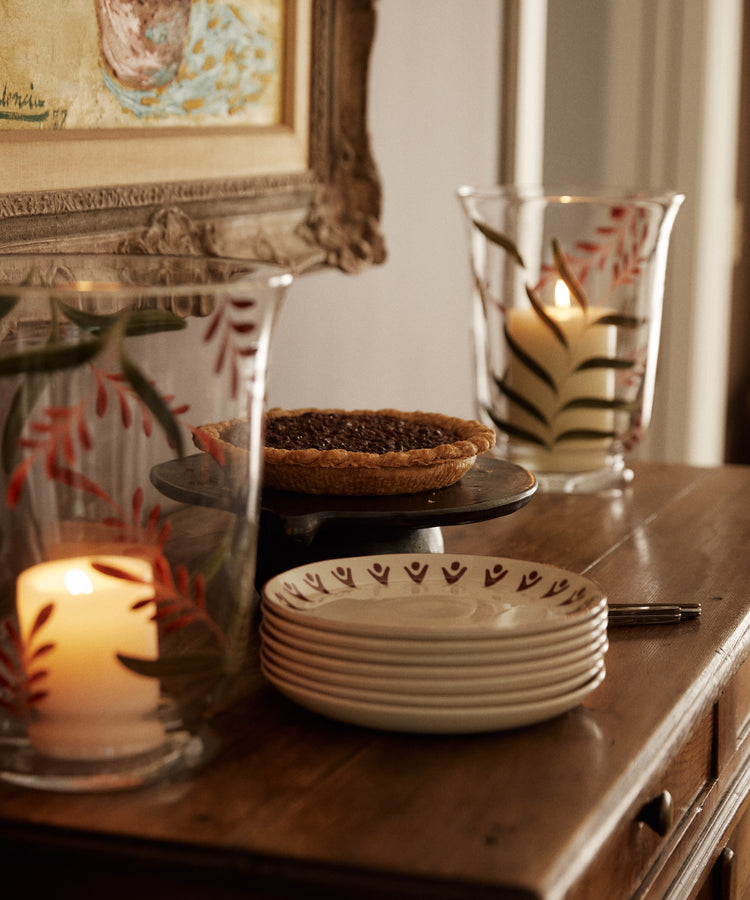 A pecan pie on a cake stand, Vito Small Plate, Set of 4, a fork, and glass candle holders with leaf designs sit on a wooden sideboard.