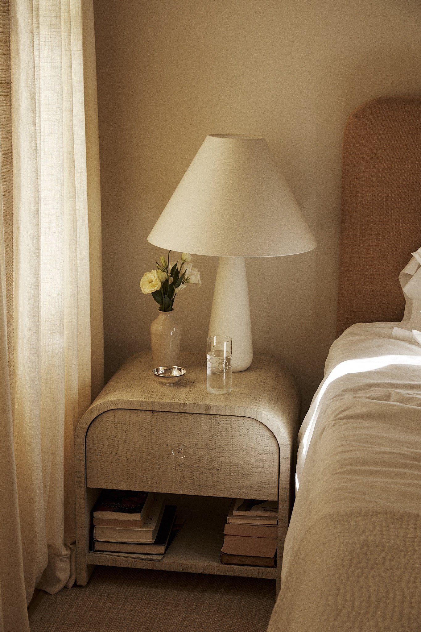 A bedside table next to a bed with white linens holds a lamp, a vase of white flowers, the Deco Tall Water Glass, Set of 6 in cobalt blue, and a small dish, with books stacked on the lower shelf.