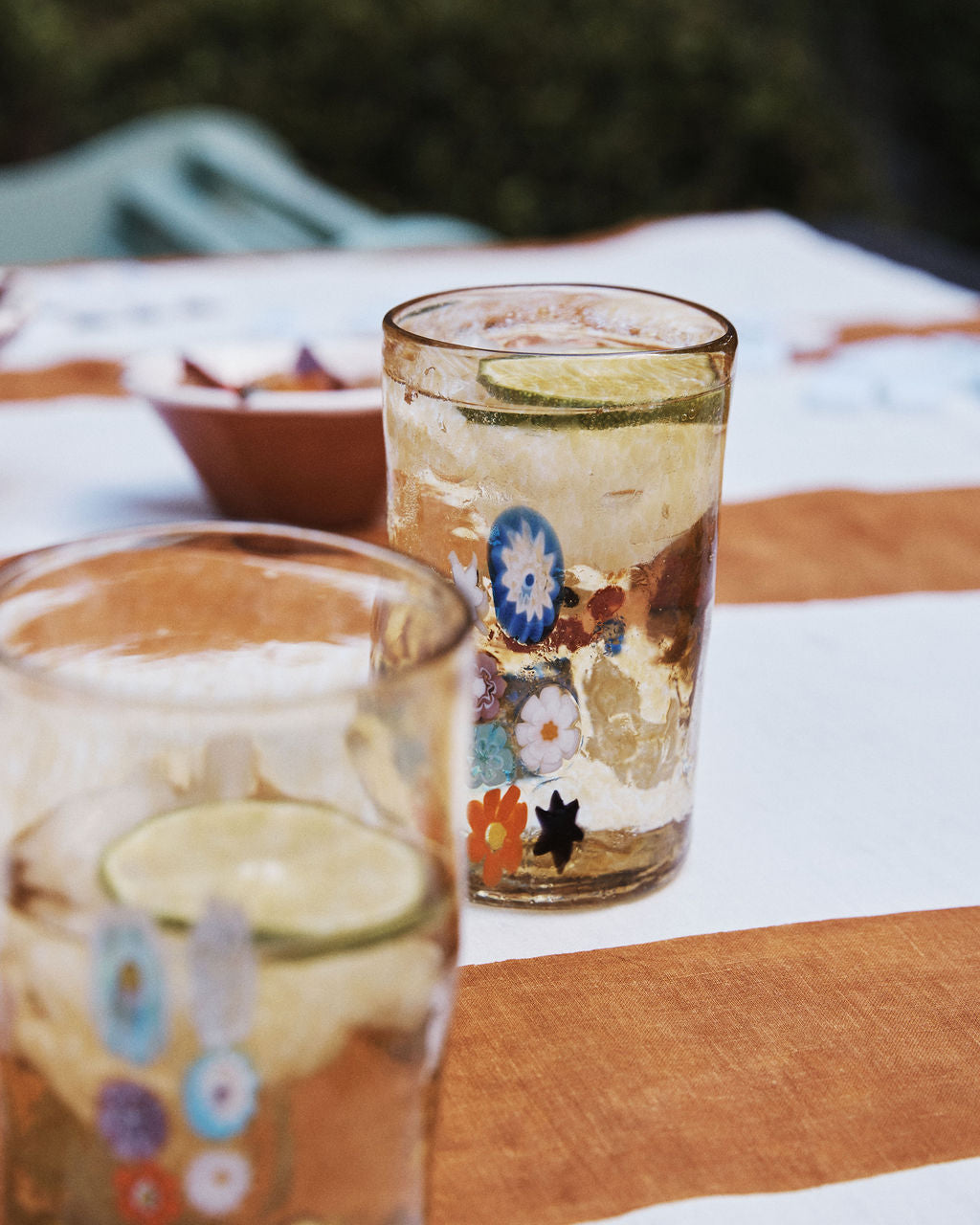 The Muranesi Glass Set of 2, featuring handblown floral designs, is shown filled with a light drink and lime slices atop a brown and white striped tablecloth.