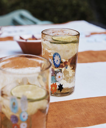 The Muranesi Glass Set of 2, featuring handblown floral designs, is shown filled with a light drink and lime slices atop a brown and white striped tablecloth.