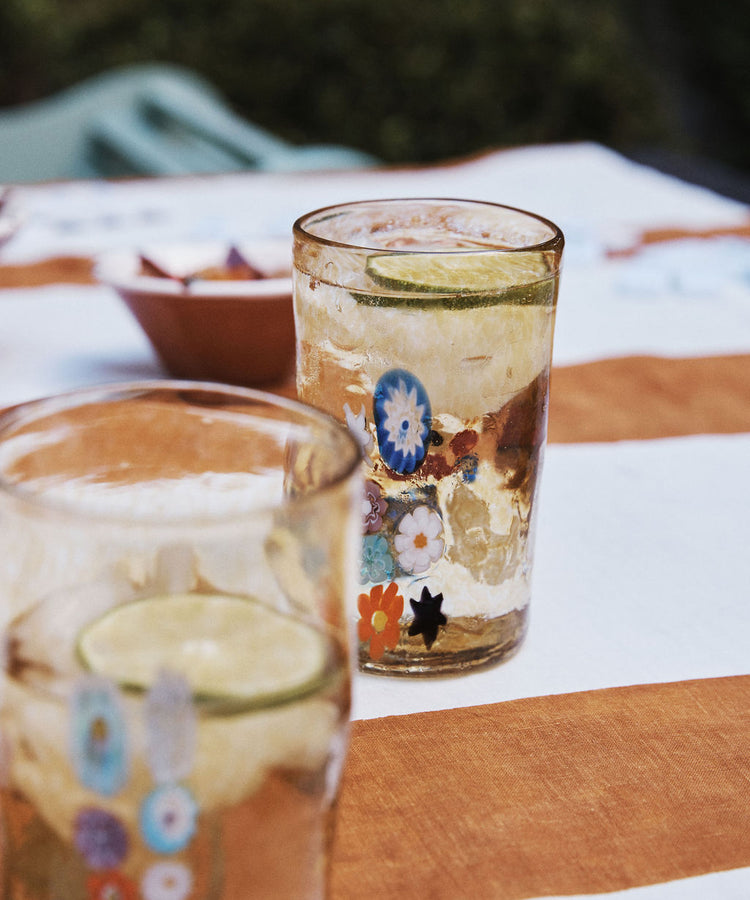 The Muranesi Glass Set of 2, featuring handblown floral designs, is shown filled with a light drink and lime slices atop a brown and white striped tablecloth.